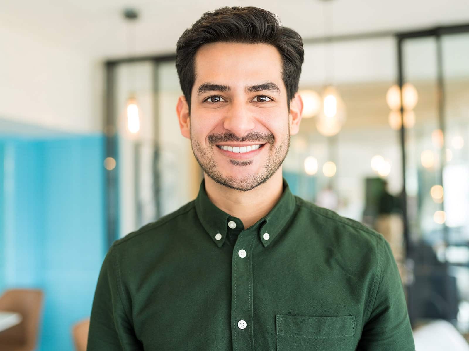 A man with dark hair and a beard smiles confidently indoors.