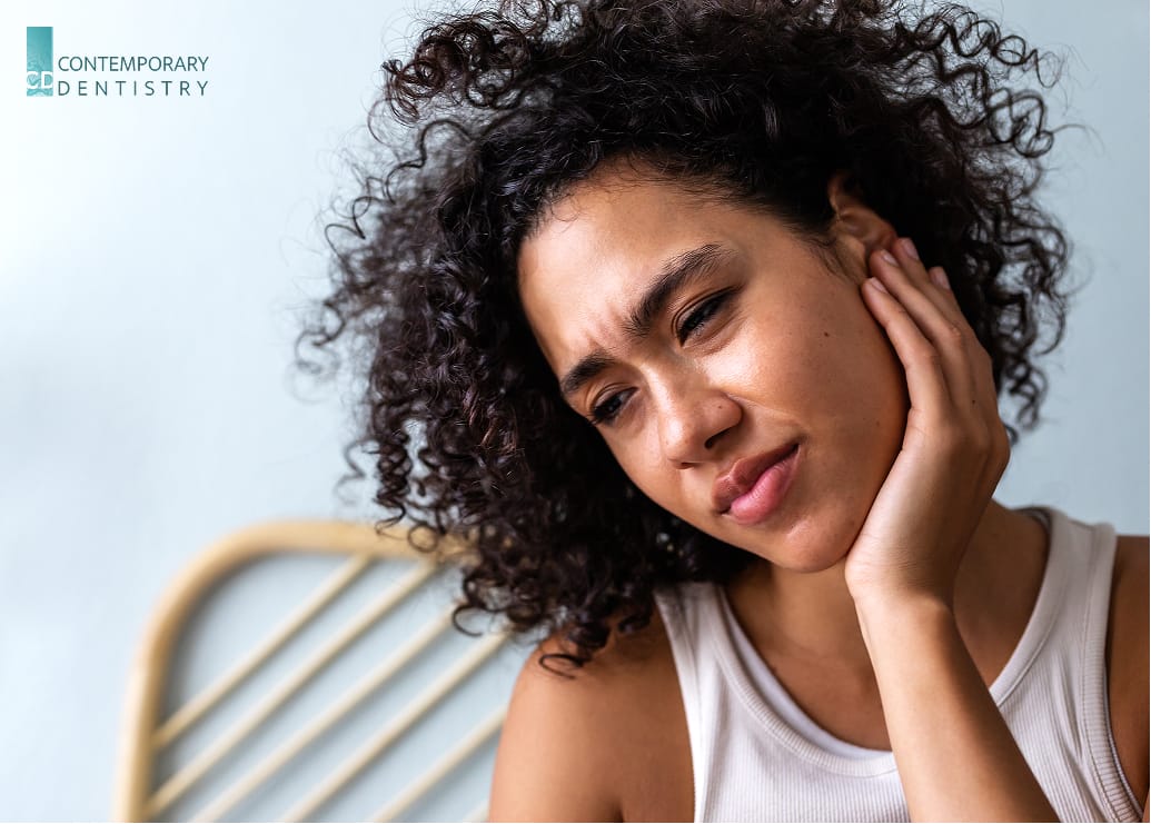 Alt text: A woman is holding her jaw with one hand, showing a distressed expression, likely due to the pain associated with TMJ (temporomandibular joint) disorder. Her other hand is placed on her cheek, suggesting discomfort or tension in the jaw area.