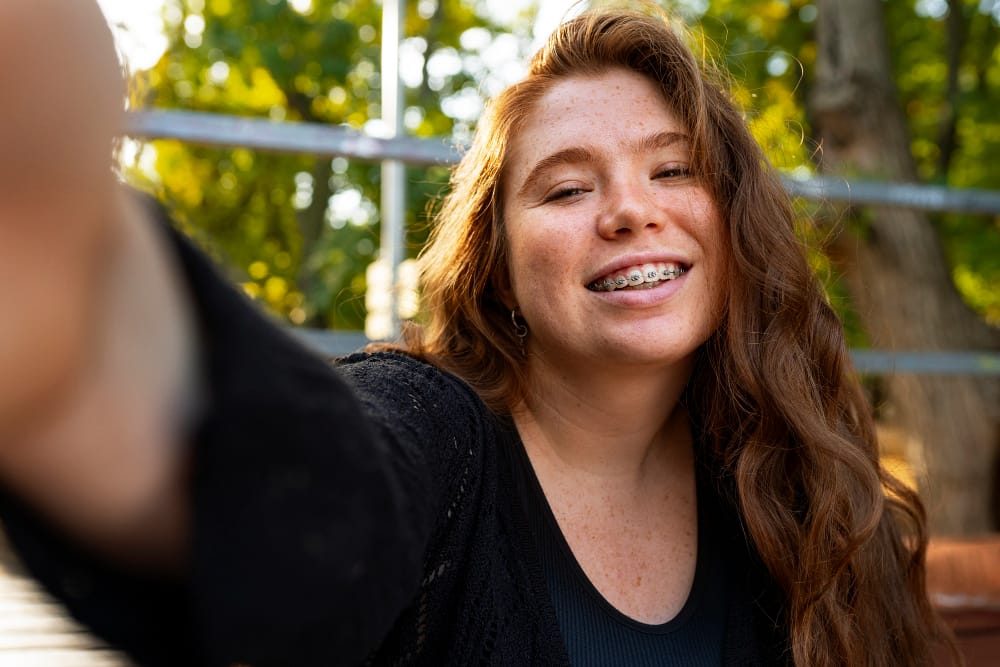 A young woman with long red hair smiles outdoors while wearing metal braces.